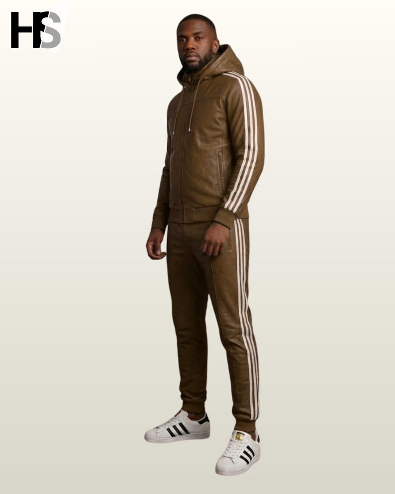 May include: A brown leather tracksuit featuring a hooded zip-up jacket and matching trousers. Both pieces are accented with three white vertical stripes along the sleeves and legs. The outfit is paired with white trainers featuring black stripes.