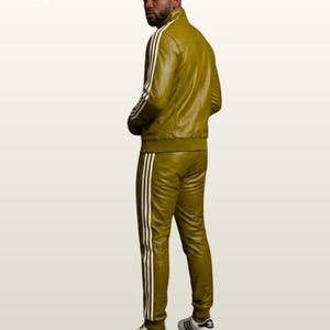 May include: An olive green leather tracksuit featuring a high-neck jacket and matching trousers, both adorned with three white vertical stripes along the sleeves and legs. The set is paired with white sneakers featuring black stripes and a logo.