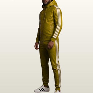 May include: An olive green leather tracksuit featuring a hooded zip-up jacket and matching joggers. Both pieces are accented with three white vertical stripes along the sleeves and legs. The outfit is paired with white sneakers with black stripes.