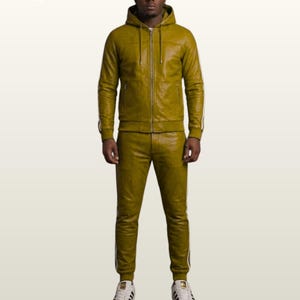 May include: An olive green leather tracksuit consisting of a zip-up hooded jacket with drawstrings and matching slim-fit joggers. Both pieces feature white side stripes along the arms and legs. The outfit is paired with white sneakers.
