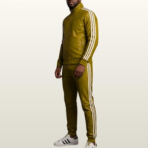 May include: An olive green track suit featuring a full-zip jacket with a high collar and matching slim-fit track pants. Both pieces are adorned with three white vertical stripes running down the sleeves and legs. The outfit is paired with white sneakers.
