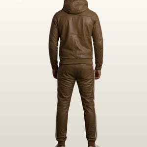 May include: A brown leather tracksuit consisting of a hooded jacket and matching trousers. The jacket features a hood and long sleeves with ribbed cuffs. The trousers have a straight-leg fit with white side stripes. White trainers are worn with the set.