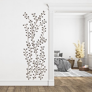 May include: A brown wall decal featuring a stylised design of leafy branches with small leaves. The decal is applied to a white wall in a room with a doorway leading to another room.