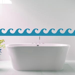 May include: A white bathtub is centered in a bathroom with a blue wave decal on the wall. A potted plant with green and yellow flowers sits to the left of the tub. The floor and walls are white.
