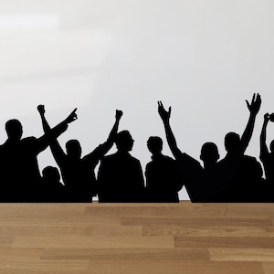 May include: Black silhouette of a crowd of people with their arms raised in the air, as if cheering. The silhouettes are cut out and placed on a white wall.