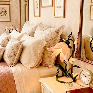 May include: A flower-shaped lamp with soft pink petals and a dark green stem sits on a white nightstand next to a round analog clock and a brown notebook. The lamp features a curved, vine-like base and emits a warm, gentle glow from its petals.