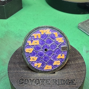 May include: A watch face with a purple honeycomb pattern and orange numerals. The watch face is set in a dark brown wooden holder. The text "COYOTE RIDGE" is visible on the holder.