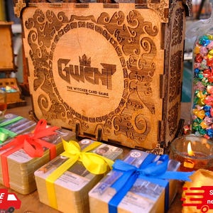 Puede incluir: Una caja de madera con el texto "Gwent: The Witcher Card Game" est&aacute; rodeada de montones de cartas atadas con cintas de colores. Tambi&eacute;n hay un tarro de cristal lleno de estrellas de origami y velas encendidas.