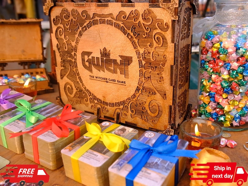 Puede incluir: Una caja de madera con el texto "Gwent: The Witcher Card Game" est&aacute; rodeada de montones de cartas atadas con cintas de colores. Tambi&eacute;n hay un tarro de cristal lleno de estrellas de origami y velas encendidas.