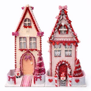 May include: Two decorative gingerbread houses with glitter and embellishments. One is light pink and the other is off-white. Both have red and pink accents, including bows, beads, and miniature Christmas trees. They sit on a white base with a fence.