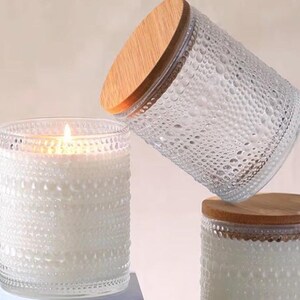 Op de afbeelding: Witte kaarsen in transparante glazen potten met een gestructureerd, bubbelachtig patroon. Elke pot heeft een houten deksel. Eén kaars is aangestoken. Decoratieve woonaccessoires.