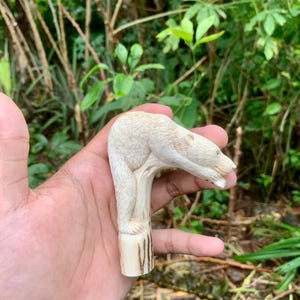 Pode incluir: Uma estatueta esculpida na cor creme representando um urso em pé sobre um tronco vertical. O urso está posicionado horizontalmente com a cabeça estendida para a frente, exibindo a textura detalhada do pelo e os traços faciais esculpidos.