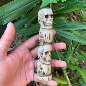 May include: A vertical totem-style carving featuring three stacked human skulls in an off-white, bone-like material. The sculpture displays detailed anatomical features including eye sockets, nasal cavities, and teeth on each individual skull.