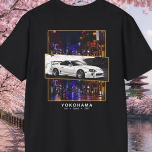 Może przedstawiać: Czarna koszulka z grafiką białego samochodu sportowego na tle nocnej scenerii miejskiej. Projekt zawiera tekst "YOKOHAMA", "A80 Supra 1993". Grafika jest stylizowaną ilustracją.