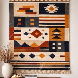 May include: A rectangular wall hanging with a geometric pattern in shades of beige, navy, rust, and gold. The design features blocks, diamonds, and triangles. The bottom edge has a fringe. A wooden rod holds the hanging.
