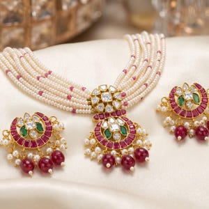 May include: A multi-strand pearl necklace featuring small pink bead accents and a central gold-tone pendant with red and green stones, clear crystals, and dangling red beads, accompanied by a pair of matching crescent-shaped stud earrings.