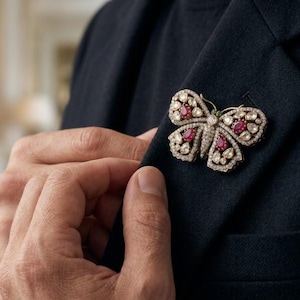 May include: A butterfly-shaped brooch crafted from gold-toned metal, featuring a pavé of small clear stones across the wings and body, accented with four pear-shaped red stones and two tiny green stones positioned at the top of the butterfly head.