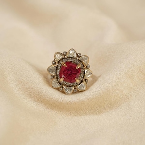 Peut inclure: Une bague ornée d'une pierre centrale rouge foncé taille coussin, entourée d'une auréole de pierres claires en forme de pétales. La monture métallique présente une finition sombre et l'ensemble forme un motif floral avec des éclats.