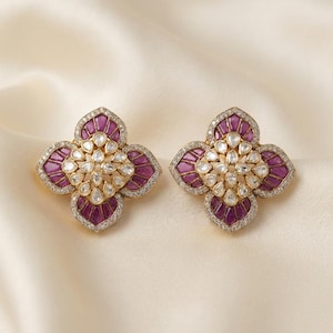 May include: A pair of flower-shaped stud earrings featuring deep magenta teardrop-shaped stones arranged in a radial pattern, accented by smaller clear teardrop stones in the center and a border of tiny white stones set in a gold-toned metal frame.