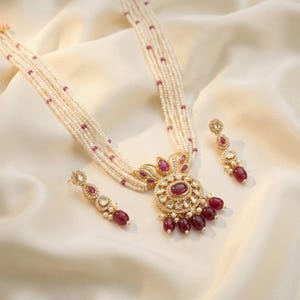 May include: A multi-strand pearl necklace featuring a central gold-toned pendant with a large oval ruby, smaller ruby accents, and clear stones. The set includes matching drop earrings with ruby stones and clear accents, all arranged in a coordinated jewelry set.