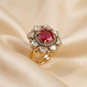 Puede incluir: Un anillo de oro con una piedra preciosa roja central de talla cojín, rodeada por un halo de pequeños diamantes redondos y un borde exterior de diamantes más grandes en forma de pera. El engaste es de oro amarillo con una banda pulida.