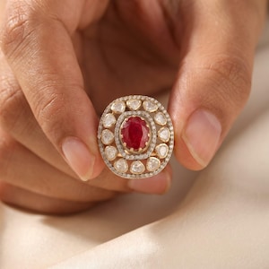 Puede incluir: Un anillo de oro con un rubí rojo central de talla ovalada, rodeado por un halo de pequeños diamantes blancos y un borde exterior de diamantes más grandes en forma de pera, engastados en una montura decorativa de varias capas.