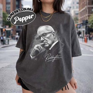 May include: A charcoal grey oversized t-shirt featuring a black and white graphic portrait of a man wearing glasses and a suit, with his finger near his mouth. The text "Reddington" is printed in white script below the portrait, and a "Comfort Colors Pepper" logo is visible in the upper left corner.