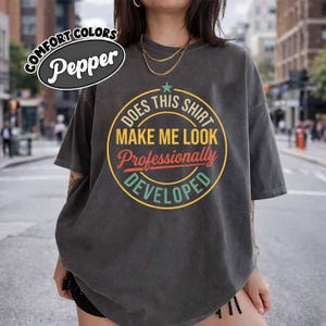 Puede incluir: Una camiseta gris carbón de manga corta con un gráfico circular y una estrella verde azulado en la parte superior. El texto dice: DOES THIS SHIRT MAKE ME LOOK Professionally DEVELOPED. La camiseta tiene un corte holgado y aspecto desgastado.
