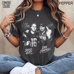 Op de afbeelding: Een antracietgrijs T-shirt met korte mouwen en een grafische print van Jared Goff en Dan Campbell. Het ontwerp bevat de tekst "Jared Goff" en "Dan Campbell" met "Lions" en het nummer "16". Het T-shirt heeft een ronde hals en opgerolde mouwen.