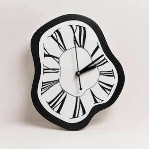 May include: A wall clock with an irregular, melting-style shape featuring a white face and a thick black border. The clock displays black Roman numerals and two black hands, with a central gold-coloured pin holding the hands in place.