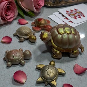 May include: A collection of decorative turtle figurines in various metallic finishes, including gold and bronze. The turtles are arranged with pink rose petals and playing cards, creating a whimsical display. The largest turtle is approximately 4 inches long.