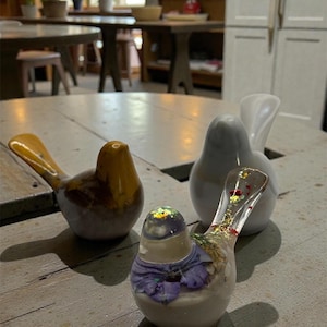 May include: A collection of decorative bird figurines. One bird is yellow and brown, two are white, and one is clear with purple floral accents and glitter. The birds are displayed on a weathered wooden table, suggesting a home decor theme.