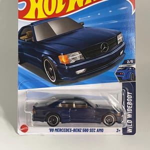 May include: A Hot Wheels die-cast model of a dark blue '89 Mercedes-Benz 560 SEC AMG. The car is displayed on a blue and white card with the Hot Wheels logo and the text "WILD WIDEBODY". The card also includes the text "GUARANTEED FOR LIFE".