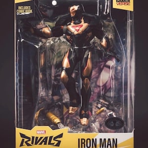 Może przedstawiać: Figurka akcji Iron Man z serii Marvel Rivals w przezroczystym plastikowym pudełku. Figurka jest czarno-złota z czerwonym, świecącym elementem na klatce piersiowej. Opakowanie zawiera napisy Marvel Rivals Iron Man, Gamer Verse oraz Includes Comic Book.