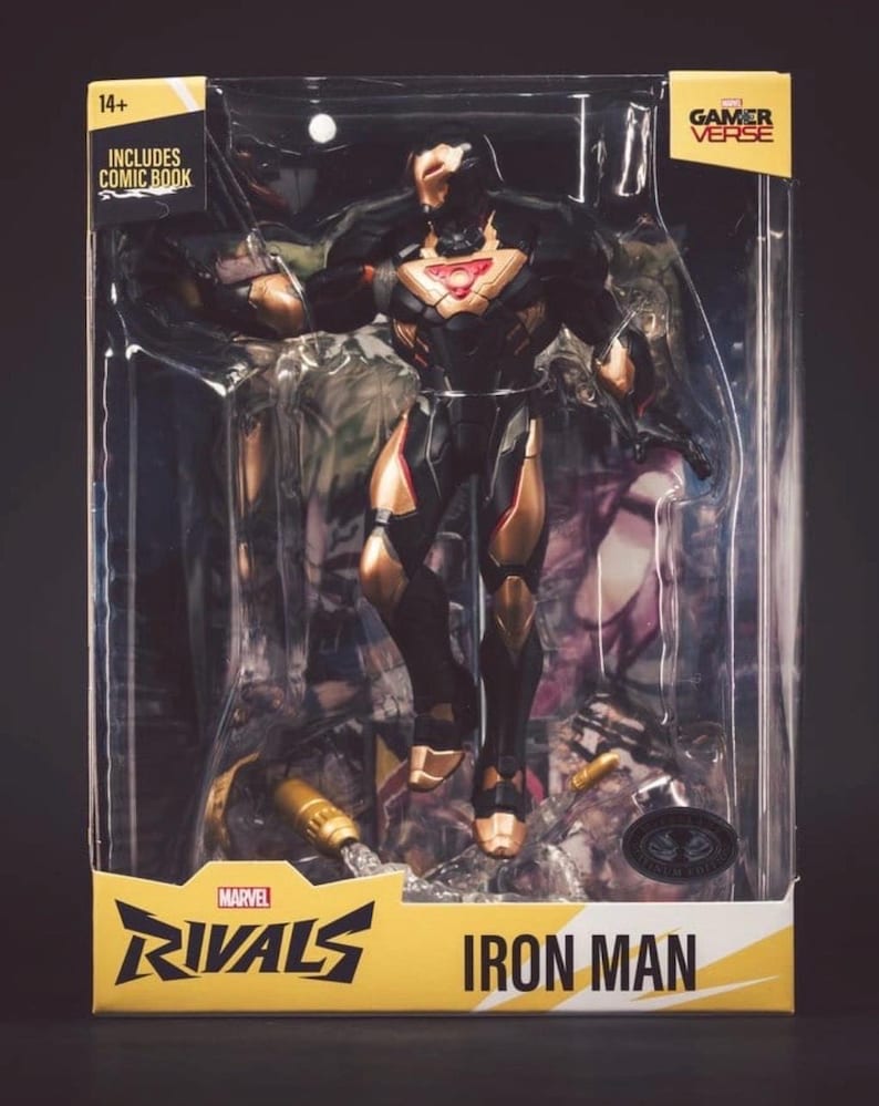 Może przedstawiać: Figurka akcji Iron Man z serii Marvel Rivals w przezroczystym plastikowym pudełku. Figurka jest czarno-złota z czerwonym, świecącym elementem na klatce piersiowej. Opakowanie zawiera napisy Marvel Rivals Iron Man, Gamer Verse oraz Includes Comic Book.