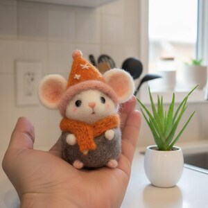 Könnte beinhalten: Eine handgefertigte Filzmausfigur mit einem orangefarbenen Hut mit Sternen, einem rosa Rand und einem orangefarbenen Schal. Die Maus hat große, runde Ohren und trägt ein graues Outfit. Die Figur wird in einer Hand gehalten.