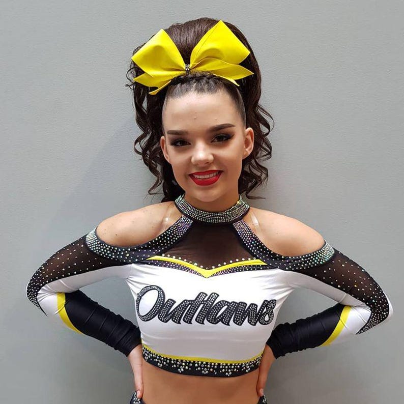 Puede incluir: Una joven con un uniforme de animadora. La parte superior blanca tiene detalles en negro y amarillo, con la palabra "Outlaws" en la parte delantera. El uniforme incluye un lazo amarillo en el pelo y un escote enjoyado.
