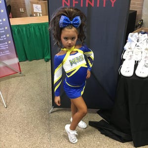 Puede incluir: Una ni&ntilde;a con un uniforme de animadora azul y blanco con la palabra "MAVS" en el pecho. Lleva un gran lazo azul en el pelo y zapatillas blancas. El fondo presenta una pancarta con la palabra "NFINITY".