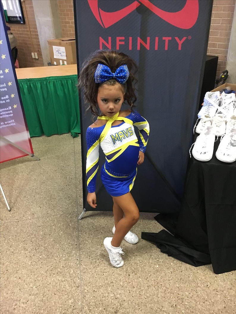 Puede incluir: Una ni&ntilde;a con un uniforme de animadora azul y blanco con la palabra "MAVS" en el pecho. Lleva un gran lazo azul en el pelo y zapatillas blancas. El fondo presenta una pancarta con la palabra "NFINITY".