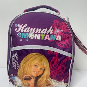 Peut inclure: Sac à déjeuner violet avec l'inscription "Hannah Montana" et une photo d'une femme blonde. Le sac a une poignée et une bande argentée. Des motifs floraux et papillons roses décorent le sac.