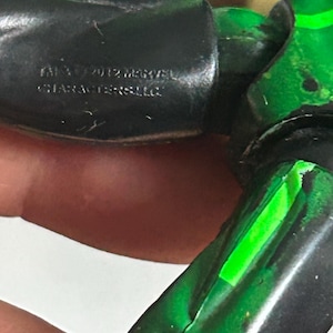 May include: A black and green articulated action figure limb featuring a molded inscription that reads 2012 MARVEL CHARACTERS LLC. The plastic surface shows visible joints and a vibrant green accent stripe running along the length of the limb.