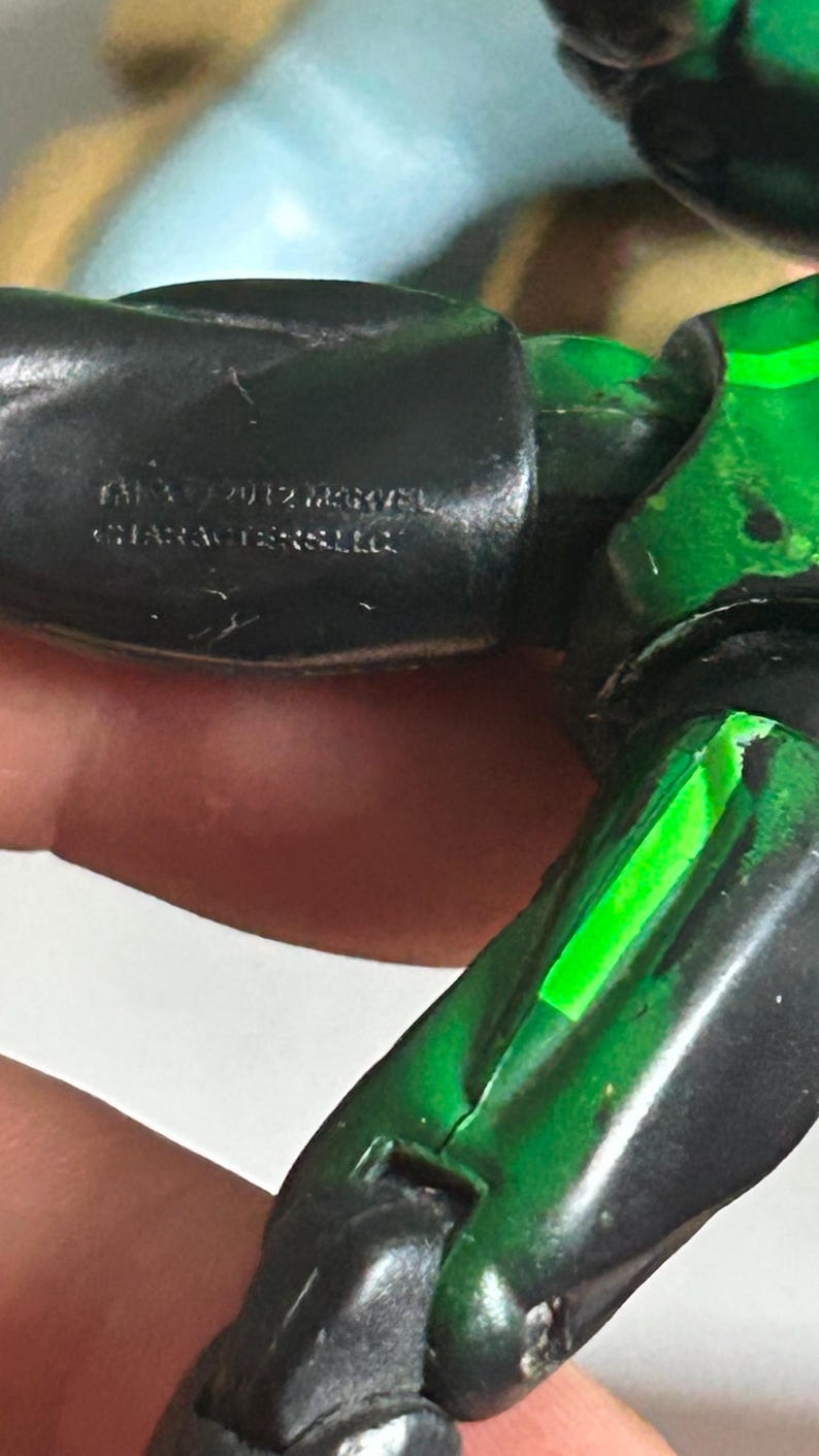 May include: A black and green articulated action figure limb featuring a molded inscription that reads 2012 MARVEL CHARACTERS LLC. The plastic surface shows visible joints and a vibrant green accent stripe running along the length of the limb.