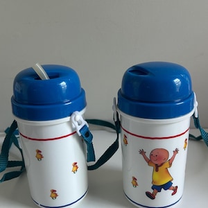 Peut inclure: Deux gourdes en plastique blanc avec des couvercles bleus à visser et des sangles de transport bleu sarcelle. Les bouteilles présentent un dessin animé d'un personnage en chemise jaune et short bleu, avec de petits oiseaux jaunes et des lignes rouges.