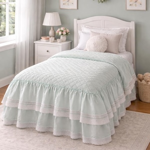 May include: A bed with a white headboard, pillows, and a light blue quilted bedspread. The bed skirt has layered ruffles with white lace trim. A white bedside table with a lamp and flowers is next to the bed.