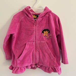 Peut inclure: Veste à capuche rose vif avec fermeture éclair et patch brodé Dora l'exploratrice. La veste a une poche avant et un ourlet à volants. Elle est suspendue à un cintre blanc.