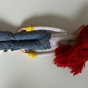 May include: A doll with bright red yarn hair, wearing a white long-sleeved shirt with yellow cuffs, blue denim-textured pants, and cow-print chaps. The doll has light-toned skin and is positioned horizontally against a plain white background.