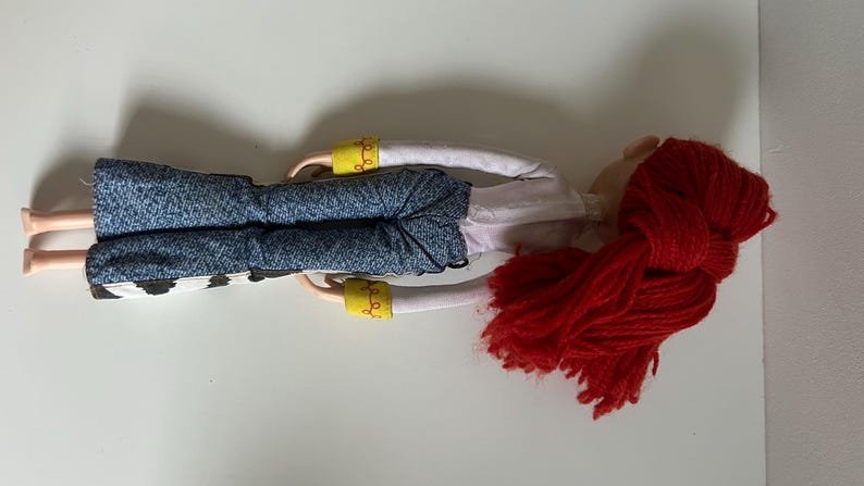 May include: A doll with bright red yarn hair, wearing a white long-sleeved shirt with yellow cuffs, blue denim-textured pants, and cow-print chaps. The doll has light-toned skin and is positioned horizontally against a plain white background.
