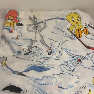 May include: White bed sheet with cartoon characters Bugs Bunny, Tweety Bird, and the Road Runner playing hockey. The sheet has blue and red accents, with the text "© WARNER BROS INC. 1980".