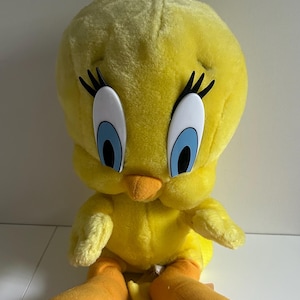 May include: A plush Tweety Bird backpack featuring a bright yellow soft fabric body, large white eyes with blue irises, long black eyelashes, and an orange beak. The character has orange feet and a yellow fabric loop handle at the top of the head.