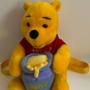 May include: A plush yellow bear backpack wearing a red shirt with the word Pooh embroidered in gold. The bear holds a blue honey pot with the word HUNNY embroidered in yellow, featuring yellow plush honey dripping over the rim of the pot.